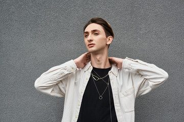 Wall Mural - A young queer person confidently poses against a gray wall, showcasing his stylish attire with pride and confidence.