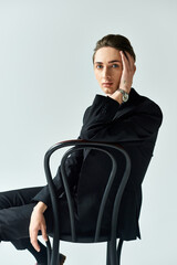 Wall Mural - A young queer person exuding confidence, sitting on a chair in a stylish black suit against a grey studio background.