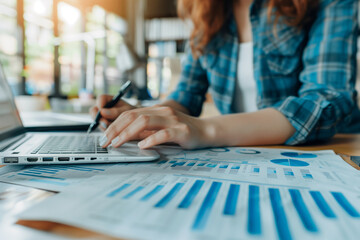 Poster - A woman is typing on a laptop while looking at a piece of paper with graphs on it. She is focused on her work and seems to be in a serious mood