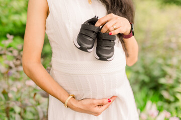 Wall Mural - A pregnant woman holds a pair of black tennis shoes over her belly. She wears a white dress. 