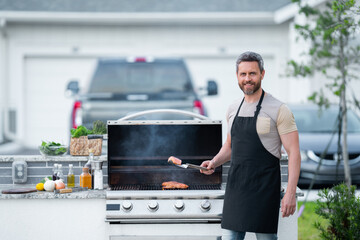 Wall Mural - Male chef grilling and barbequing in garden. Barbecue outdoor garden party. Handsome man preparing barbecue meat. Concept of eating and cooking outdoor during summer time.