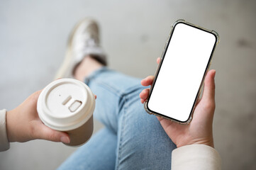 Wall Mural - A close-up image of a woman holding a takeaway coffee cup and a smartphone, sitting indoors.