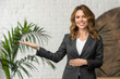 © BetterPhoto - Smiling businesswoman in a suit presenting with a welcoming gesture in a modern office