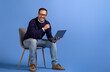 © Moon Safari - Successful male manager with laptop smiling at camera while sitting on chair over blue background