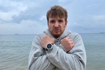 Young man shivering and trembling from cold rainy weather at summer vacation on beach, sea