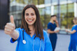 © Dragana Gordic - A joyful Caucasian female nurse gestures positively with a thumbs up while wearing blue scrubs, a stethoscope around her neck, standing before a hospital with colleagues in the background.