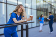 © Dragana Gordic - A female healthcare worker in blue scrubs appears stressed during a break, holding a coffee cup outside a medical facility, as her colleagues engage behind her.