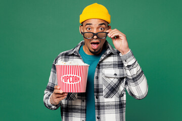 Wall Mural - Young shocked surprised amazed man wear shirt blue t-shirt yellow hat lower 3d glasses watch movie film hold bucket of popcorn in cinema look camera isolated on plain green background studio portrait.