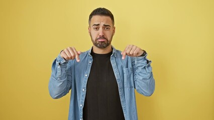 Poster - Upset young hispanic man in denim shirt pointing down, displaying a mix of unhappy, sad, and depressed emotions. isolated against a vivid yellow background.