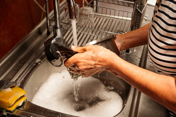 A woman washes a dirty dish in a sink. The sink is full of soapy water and the woman is using her hands to scrub the dish