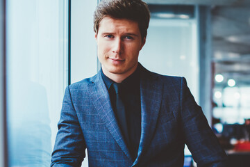 Portrait of confident businessman in elegant suit standing near office window and looking at camera, handsome caucasian male entrepreneur spending time at coworking space and waiting colleague