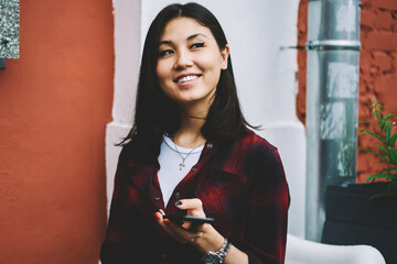 Wall Mural - Young asian female student checking notifications via mobile phone while looking away with smile on face outdoors, cheerful trendy hipster girl using telephone app for searching some cafeteria