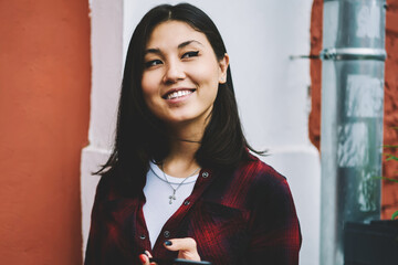 Wall Mural - Young asian female student checking notifications via mobile phone while looking away with smile on face outdoors, cheerful trendy hipster girl using telephone app for searching some cafeteria