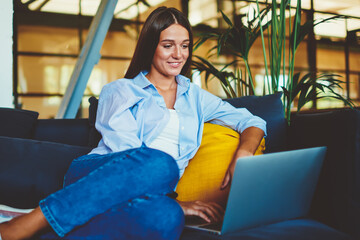 Wall Mural - Positive smiling hipster girl searching comedy show for watching using modern laptop device indoors, happy woman resting on comfortable sofa while messaging with friends via application on netbook