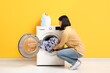 © Pixel-Shot - Young woman putting dirty clothes into washing machine on orange background