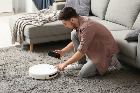 Young man with mobile phone turning on modern robot vacuum cleaner at home