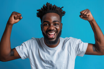Wall Mural - Photo of an excited black man wearing a casual t-shirt while raising fists up on a blue background