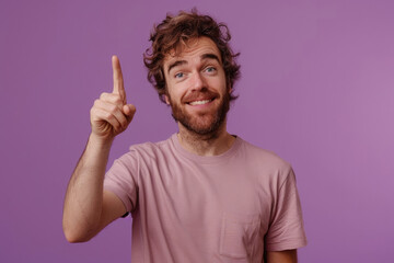 Wall Mural - Photo of an enthusiastic man with a thin beard wearing a casual t-shirt while raising his index finger up against a purple background