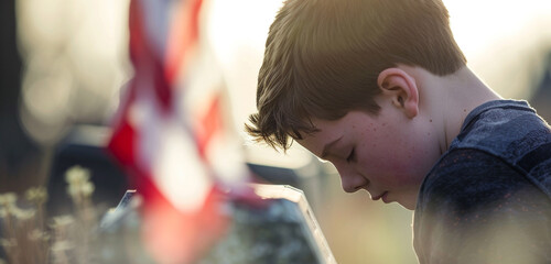 Wall Mural - boy in deep thought at a tombstone, head bowed with a blurred us flag behind.