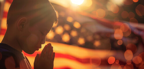 Wall Mural - boy in sharp focus praying with a blurred us flag behind in a bokeh effect, creating a high-contrast