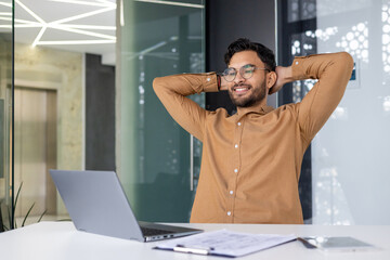 Poster - Young professional relaxing with hands behind head at modern office desk with laptop