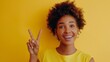 © DigitalParadise - Portrait of beautiful young black woman in yellow top with short curly hair, smiling and making the peace sign on a yellow background, template for advertising or promotion, with copy space for text