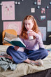 © AnnaStills - Vertical shot of modern teen girl with red hair drinking milk tea while reading book in her bedroom