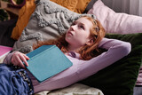 Fototapeta  - Teen girl holding book relaxing on bed in her bedroom thinking about plot of novel she reading, copy space