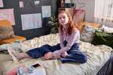Fototapeta  - Red-haired teen girl wearing casual clothes sitting on bed in her bedroom at home