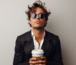 © AmayaGB - Hombre joven 40s, tez morena, gafas de sol cabello rizado despeinado camisa blanca traje, sujetando un vaso de café sobre fondo claro, de frente, recurso gráfico, seguridad, finanzas, trabajo se busca