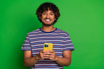 Sticker - Portrait of young curly haired guy in striped t shirt holding modern smartphone eshopping features isolated on green color background