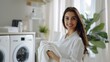 © Iona - young woman holding freshly folded laundry, smiling in a bright laundry room. The room is well-organized and filled with natural light, creating a clean and efficient atmosphere