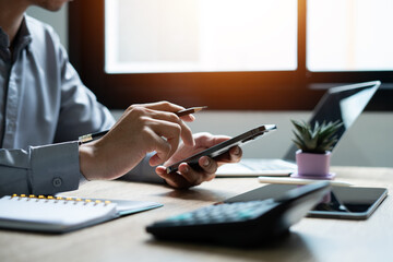 Wall Mural - A man is sitting at a desk with a laptop, a cell phone, a calculator, and a potted plant. He is using his cell phone and pen to take notes