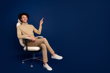 Poster - Full body photo of attractive young man sit armchair point empty space dressed stylish beige clothes isolated on dark blue color background