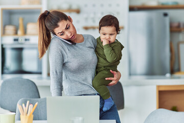 A busy mother multitasking and working from home while talking on a phone and carrying her small child