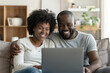 © AI_images - A happy African American middle-aged couple is using a laptop together while relaxing on the couch at home. The smiling mature man and woman look at the computer screen, watching a video, browsing, or