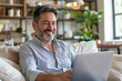 © AI_images - A happy Latino middle-aged man is using a laptop while sitting on the sofa at home. The mature older user gazes at the computer screen, browsing the internet and engaging in ecommerce shopping on a