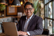 © AI_images_for_people - A Latino businessman works on his laptop in a cafe, embodying the concept of remote and freelance work. His smile radiates success as he sits at a wooden desk, bathed in sunlight. Dressed sharply in a