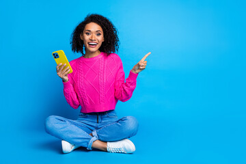 Poster - Full size photo of pretty young girl sit floor point empty space hold device wear trendy pink outfit isolated on blue color background