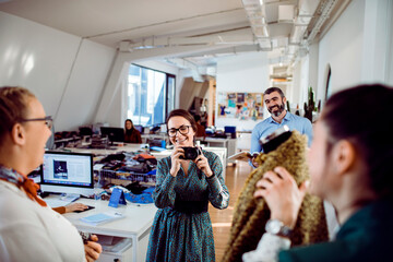 Wall Mural - Smiling female fashion designer holding a camera in a lively design studio with colleagues examining fabric in the background