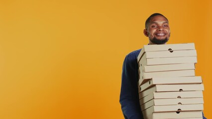 Wall Mural - Confident male courier carrying big pile of pizza boxes to deliver fast food order to clients, takeaway service in studio. Young smiling deliveryman bringing takeout meal stack. Camera B.
