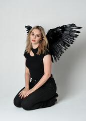 Canvas Print - Full length portrait beautiful blonde girl wearing modern black shirt, leather pants, feather angel wings, casual halloween costume. Kneeling sitting pose, isolated figure on white studio background.