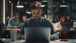 © Scott - A young professional sitting at a desk in a contemporary office space, typing on a laptop and surrounded by colleagues dressed in business casual attire.
