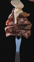 Poster - Freshly grilled pieces of beef steak rotate on a fork on a black background, pour pepper sauce. Close-up of food