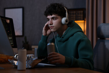 Wall Mural - Young man with energy drink and headphones playing video game at wooden desk indoors