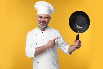 Wall Mural - Happy chef in uniform pointing at wok on orange background