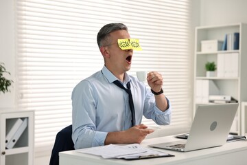 Wall Mural - Man with fake eyes painted on sticky notes holding cup of drink and yawning at workplace in office