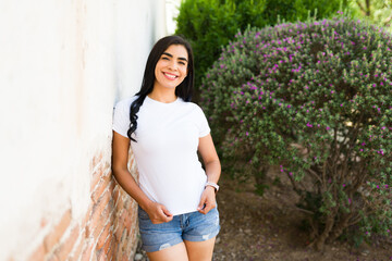 Cheerful hispanic woman in a casual white t-shirt posing with hands on hips near a garden, perfect for mockup designs
