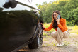 © Home-stock - Worried young European woman talking on cellphone and checking her car flat tire on the side of the highway, calling for help