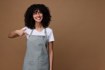 Wall Mural - Happy woman pointing at kitchen apron on brown background, space for text. Mockup for design